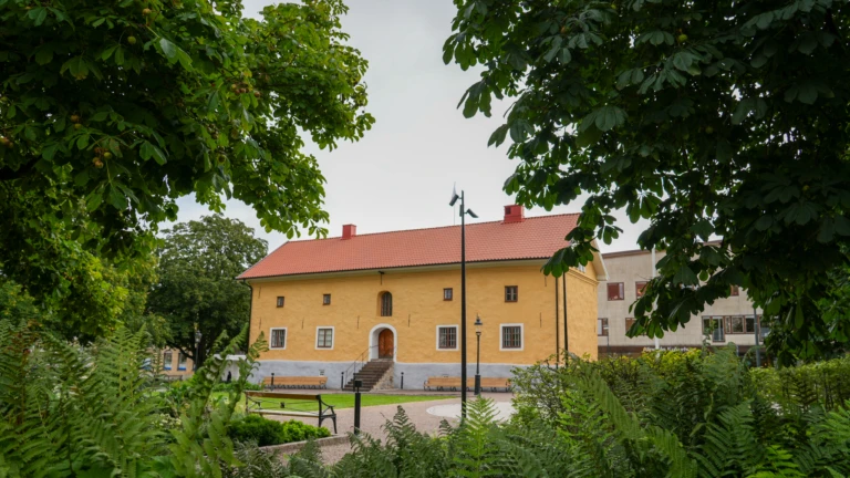 Gul museibyggnad omringad av gröna träd och växter.