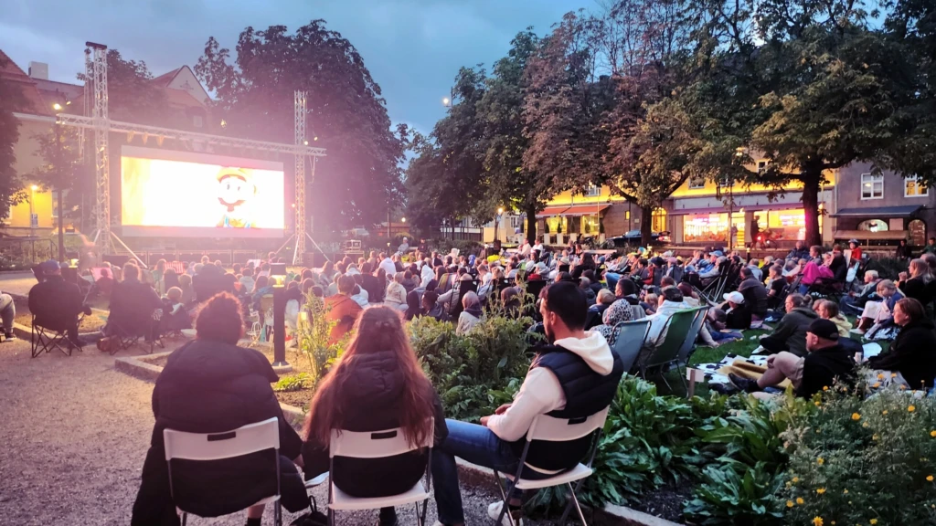 Hundratals personer sitter i en park och kollar på en stor skärm i skymning.