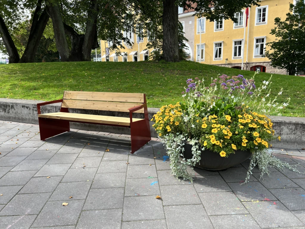 Träbänk tillsammans med en stor urna fylld med gula och vita blommor. I bakgrunden skymtar Grand Hotel Alingsås.