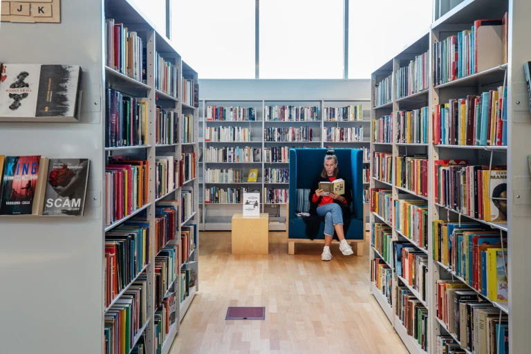 Ung kvinna sitter i en fåtölj och läser en bok i ett bibliotek.