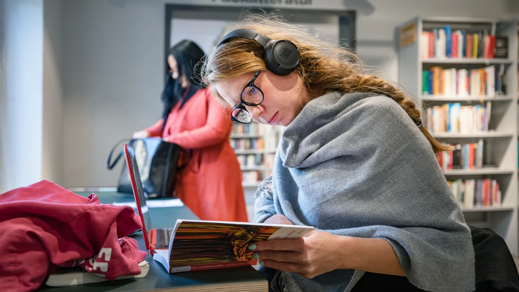 Tjej med hörlurar sitter på bibliotek och bläddrar i en bok.