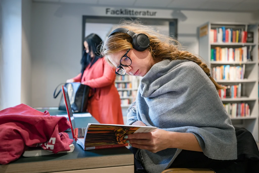 Tjej med hörlurar sitter på bibliotek och bläddrar i en bok.