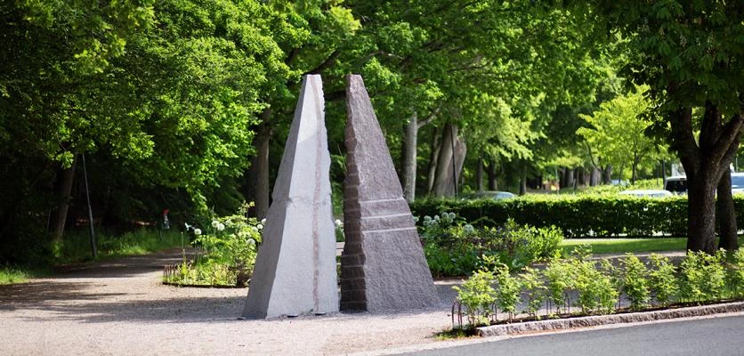 En skulptur i parkmiljö på sommaren. Två trekanter i sten står mot varandra.