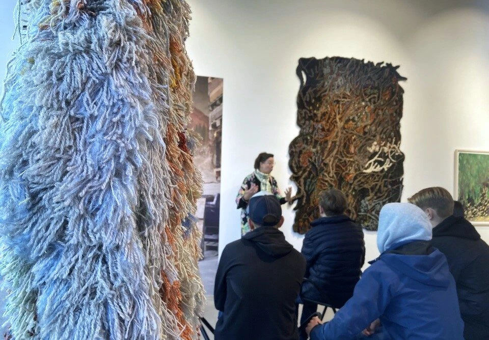 I förgrund ett konstverk i tufftat tyg. I bakgrunden sitter ungdomar. En person pratar med dem.