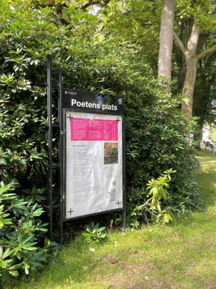 En skylt med text. Grönskande buskage i bakgrunden.
