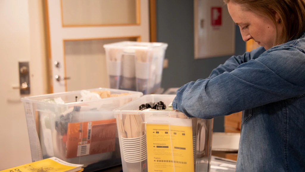 En kvinna packar transparanta plastlådor med beredskapsmateriel. I lådan finns bland annat engångsbestick, broschyren "Om krisen eller kriget kommer" samt våtservetter.