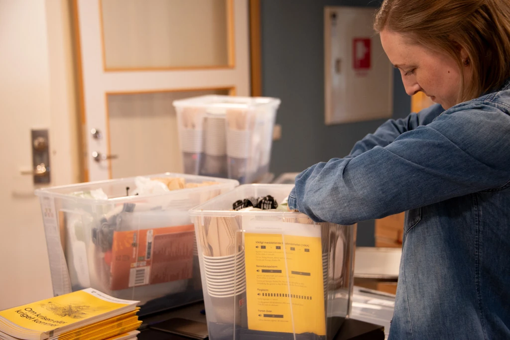 En kvinna packar transparanta plastlådor med beredskapsmateriel. I lådan finns bland annat engångsbestick, broschyren "Om krisen eller kriget kommer" samt våtservetter.