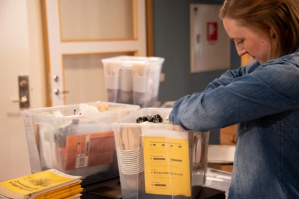 En kvinna packar transparanta plastlådor med beredskapsmateriel. I lådan finns bland annat engångsbestick, broschyren "Om krisen eller kriget kommer" samt våtservetter.