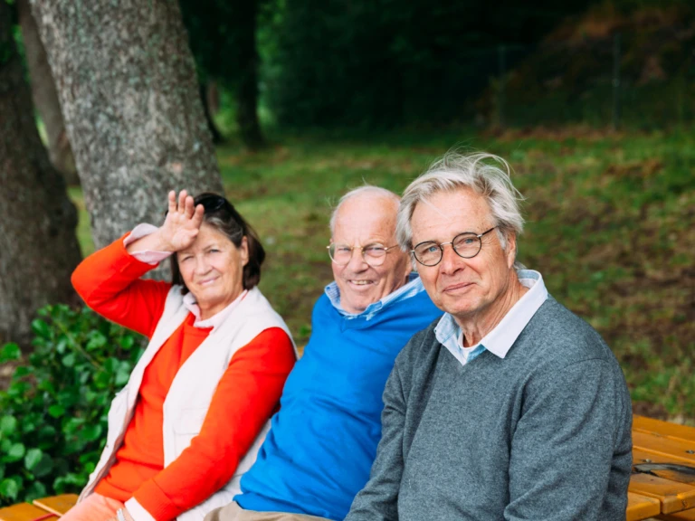 Tre äldre - en kvinna och två män sitter på en bänk och kollar in i kameran