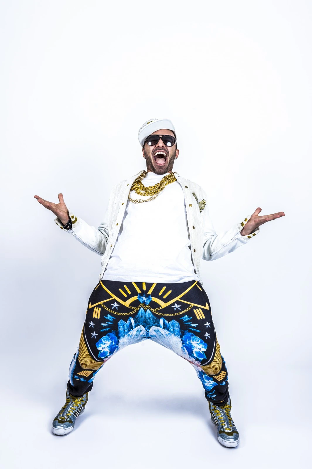 En glad man som står lite tillbakåtlutad med händerna uppe i luften vid sidan om sig. Han har keps, solglasögon, kedja runt halsen. Klädd i vit T-shirt, vit jeansjacka och pösiga byxor i olika färger.