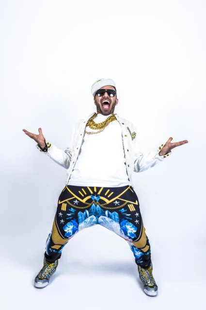 En glad man som står lite tillbakåtlutad med händerna uppe i luften vid sidan om sig. Han har keps, solglasögon, kedja runt halsen. Klädd i vit T-shirt, vit jeansjacka och pösiga byxor i olika färger.