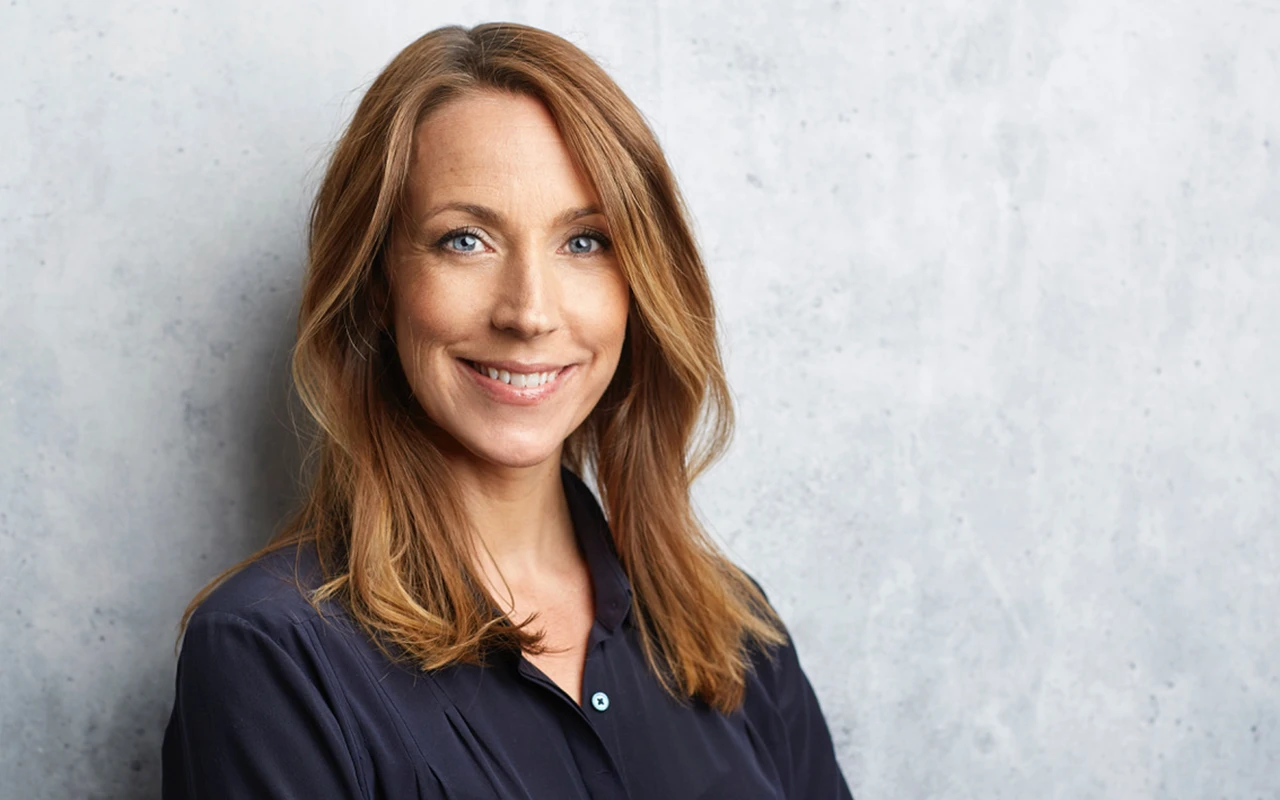 En kvinna med rött hår och svart skjorta som kollar in i kameran och ler