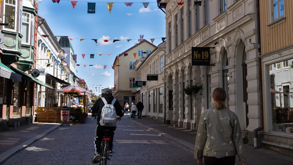 Kungsgatan i Alingsås. Vimplar hänger mellan byggnaderna. En person cyklar med ryggen mot kameran och en annan promenerar i samma riktning. Det är sol och klarblå himmel.