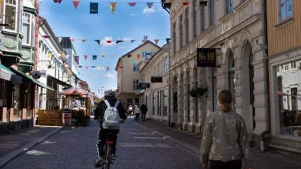 Kungsgatan i Alingsås. Vimplar hänger mellan byggnaderna. En person cyklar med ryggen mot kameran och en annan promenerar i samma riktning. Det är sol och klarblå himmel.