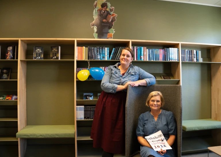 Två kvinnliga lärare i ett skolbibliotek. den ena läraren sitter i en fåtölj, den andra står lutad mot fåtöljen. båda tittar in i kameran